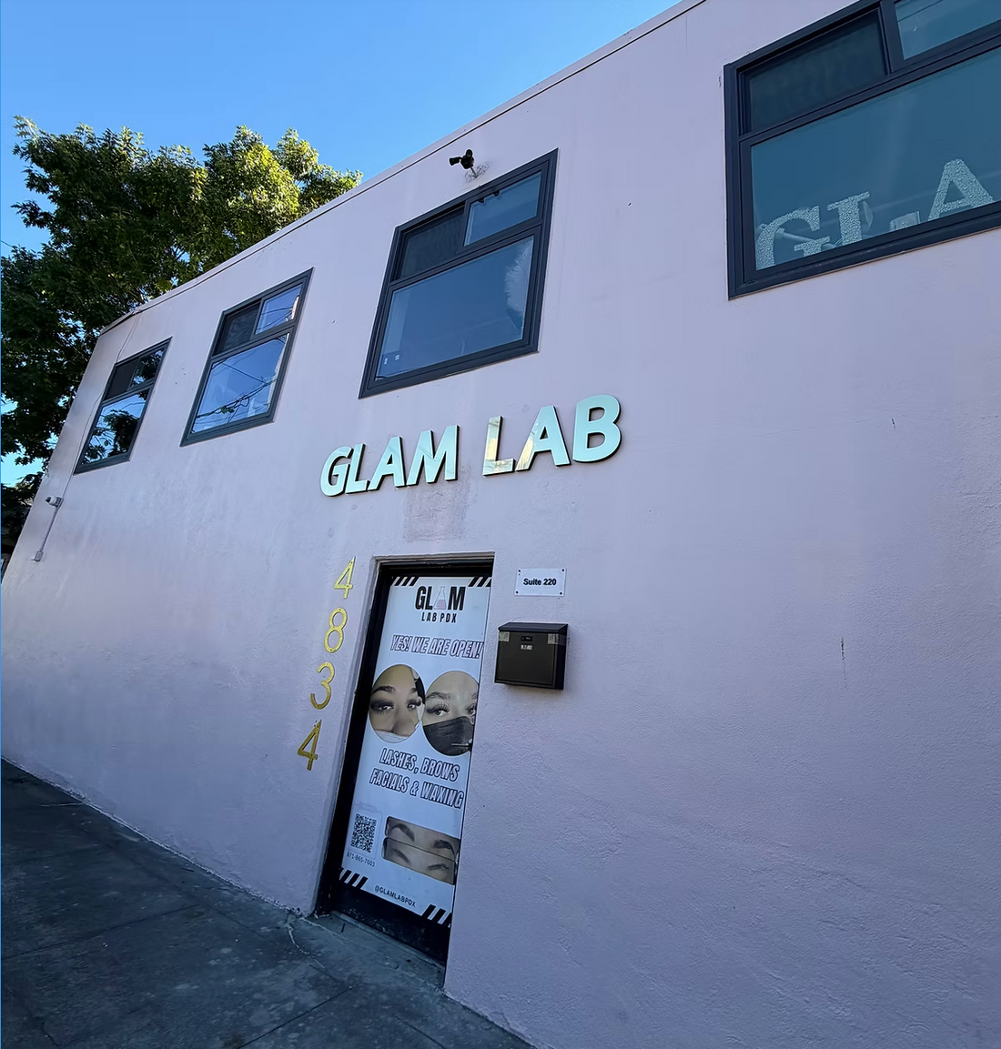 Exterior of Glam Lab PDX in Portland, Oregon, featuring a blush-pink building with teal “GLAM LAB” signage above the entrance, street-facing windows, and a branded door sign advertising lashes, brows, facials, and waxing.